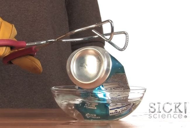 Student's gloved hand holding tongs over a crushed soda can sitting in a bowl of water (Seventh Grade Science)