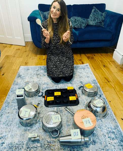 A teacher holding a wooden spoon in front of sticky notes with sight words stuck to various pots and pans and other metal objects