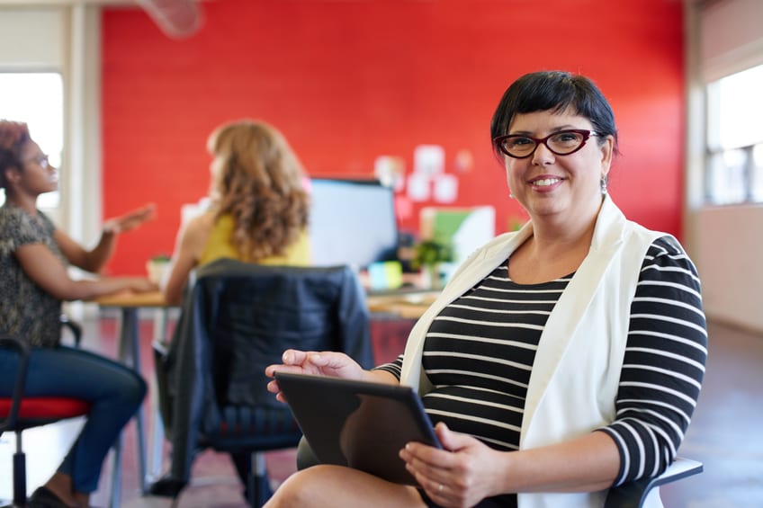 Casual portrait of a business woman using technology in a bright and sunny startup with the team in the background