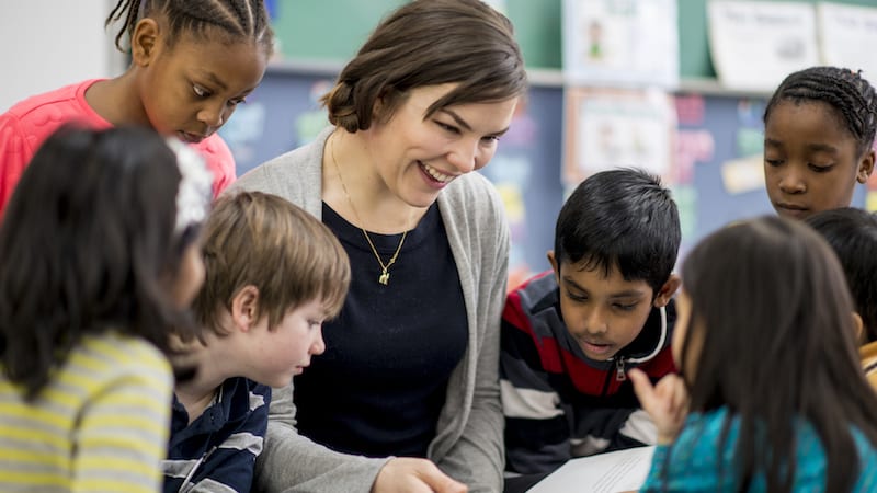 A multi-ethnic group of elementary school children are indoors in a classroom. They are wearing casual clothing. The students are gathered around their teacher to listen during story time.