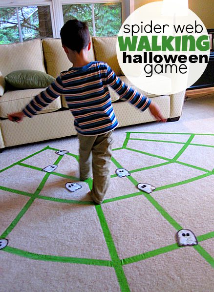 Green painter's tape is laid out on the floor in the shape of a spider web. little ghosts are scattered around it. A young boy is trying to keep his feet on the webbed lines (Halloween Activities)