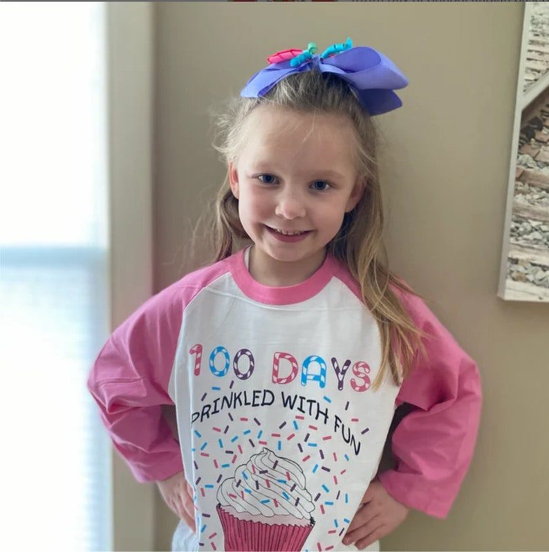 فتاة صغيرة ترتدي قميصًا بأكمام وردية اللون عليها كب كيك وتقول 100 يوم مع رش المرح. 