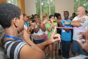 a teacher and students standing in a circle, each person with their arms crossed in front of them holding up a straw between their pointer finger and their neighbor's