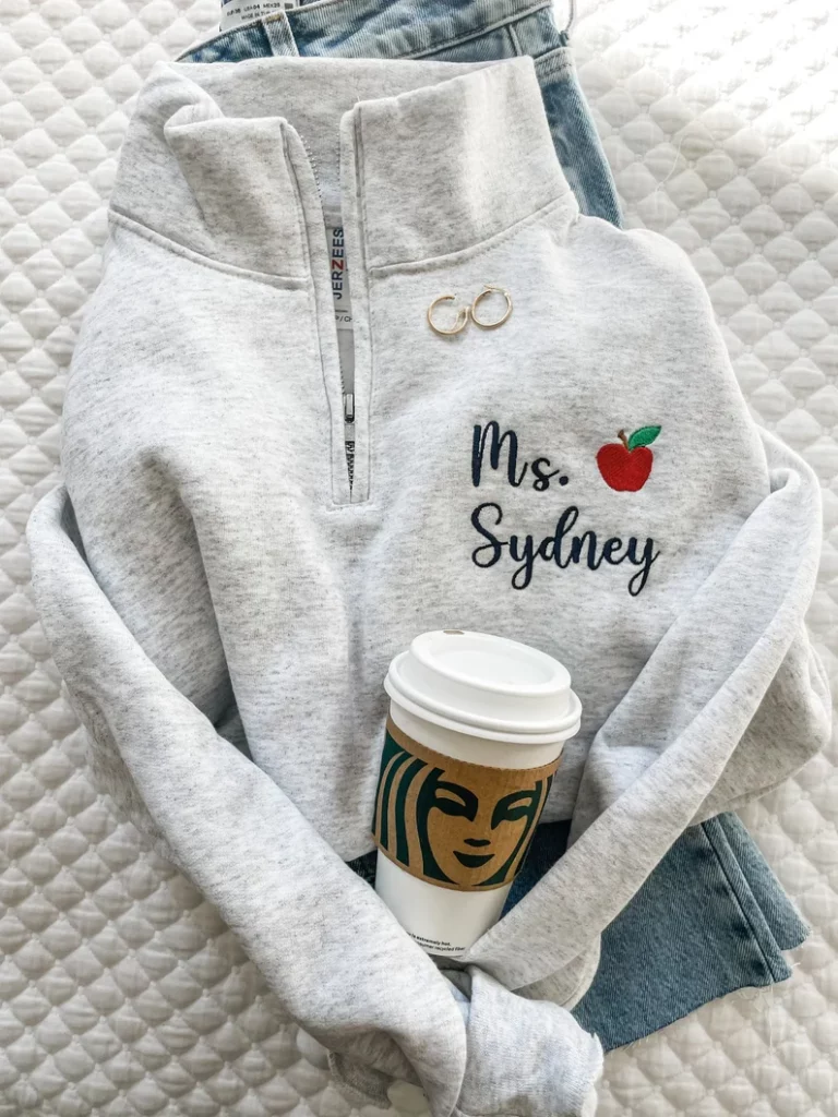 A gray quarter zip sweatshirt says Mrs. Sydney and has an apple embroidered on it. (personalized teacher gifts)