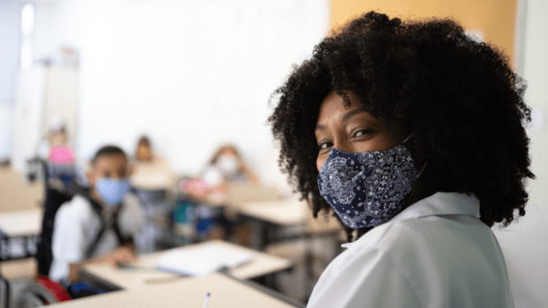 African American teacher wear mask in classroom practicing teacher self care with middle grade students wearing masks seated at desks behind her