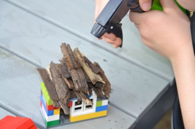 Estudiante de ciencias de tercer grado rociando agua sobre una casa de LEGO con techo de madera