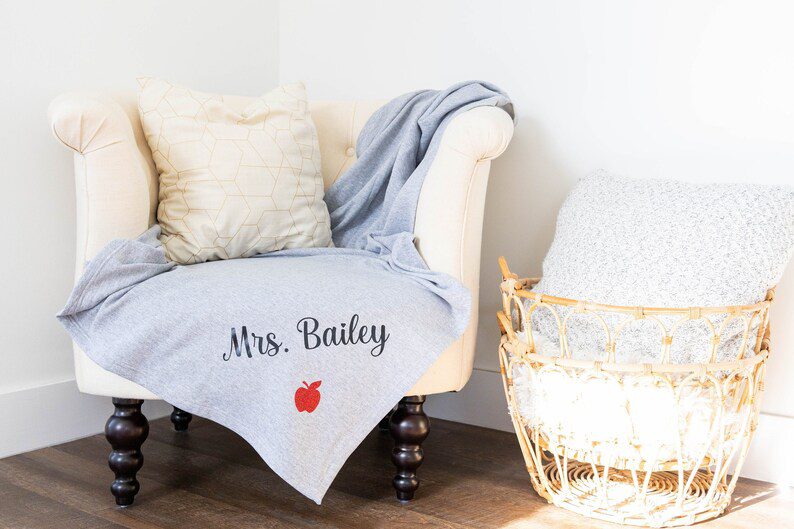 A gray fleece blanket is shown on a chair. It has a small red apple and black wording that says Mrs. Bailey.