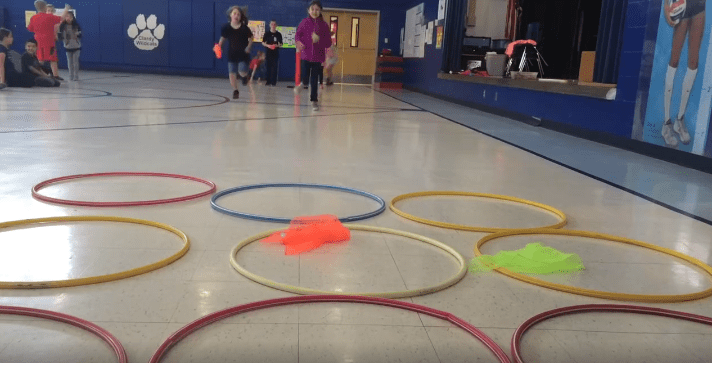 Students stand in the background. In the foreground are several hula hoops laid out on the floor (elementary PE games)