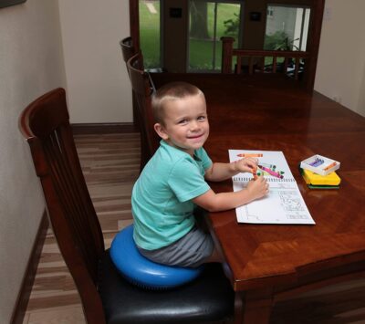 A little boy sits writing on a table while sitting on an inflatable blue cushion.