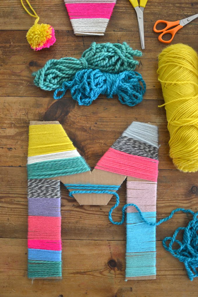 A letter M made of cardboard is wrapped with different colored yarn. Several rows of yarn are in the background.