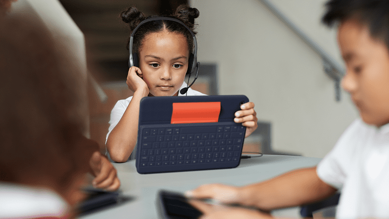 young girl sitting at table at school wearing a headset and holding a tablet, as example of tech tool ideas