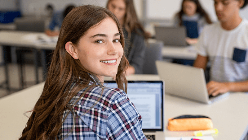 Genç kız dizüstü bilgisayarda çalışırken omzunun üzerinden kameraya gülümsüyor.